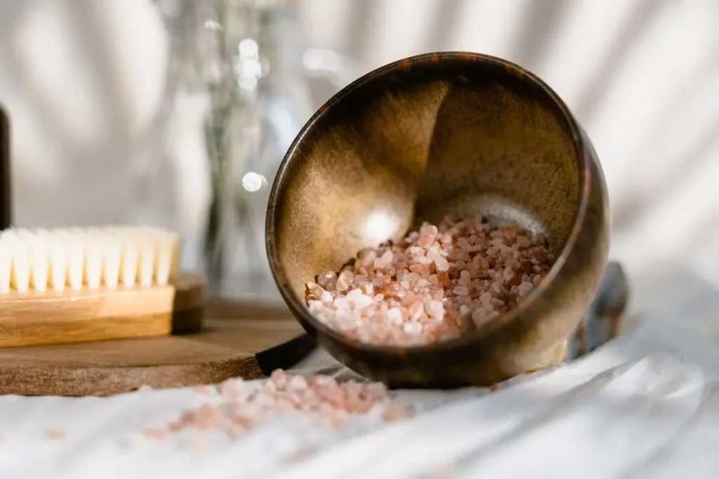 An artisanal ceramic bowl filled with coarse pink sea salt resting on a minimalist bathroom counter next to a wooden body brush.