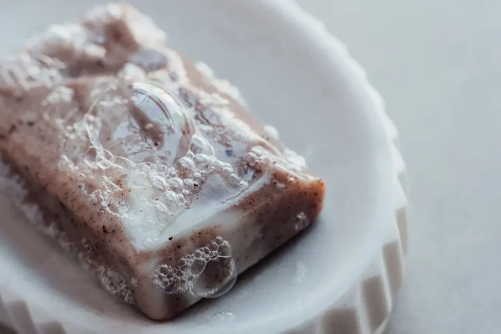 A textured handmade natural soap bar with creamy white lather bubbles resting on a minimalist ceramic soap dish.